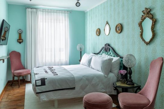 A luxurious turquoise and gold bedroom featuring a plush velvet headboard, ornate mirrors, and pink velvet accent chairs, creating a vintage-inspired boutique hotel aesthetic.