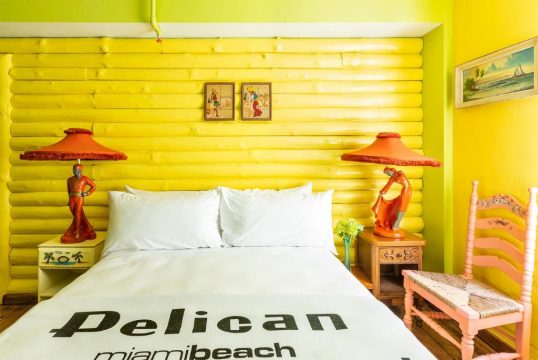 Bright yellow log walls in a hotel room with a king-size bed featuring white linens and "Pelican miamibeach" text. Two orange lamps with figurine bases flank the bed, with a pink chair to the right.