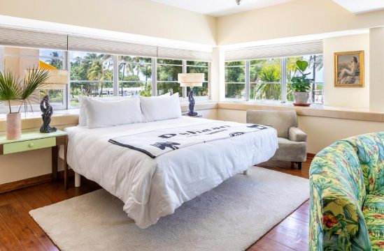 Bright bedroom with king-size bed, white linens, and "Delano Beach" throw, overlooking lush palm trees through large windows.