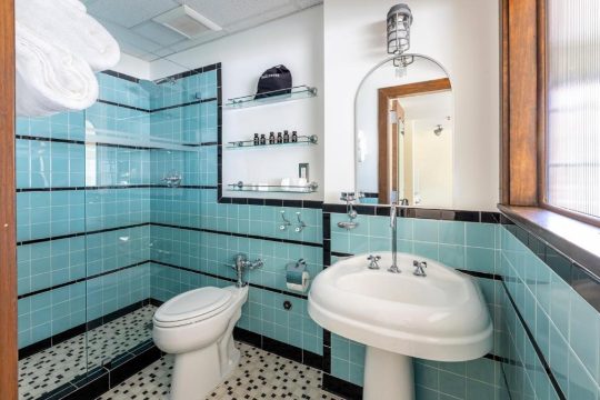 A vintage-style bathroom features a pedestal sink, toilet, and a shower with bright turquoise tiles and black trim. White towels are neatly stacked on a shelf above the toilet.