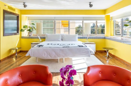 Bright yellow walls and white bedding in a sunny bedroom with orange chairs and a tropical view.