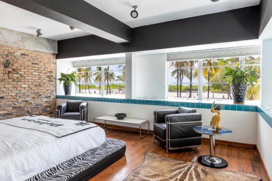 Modern bedroom with exposed brick, black leather chairs, and a king-size bed with zebra print bedding, overlooking a tropical beach with palm trees.