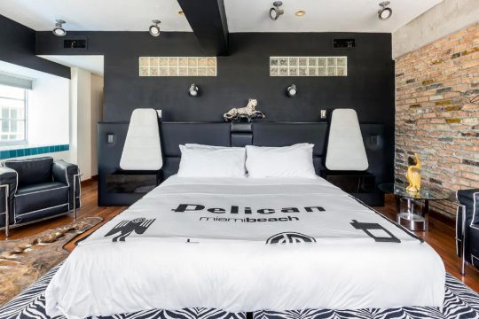 Modern black and white bedroom with a king-size bed, featuring a white duvet with "Pelican miamibeach" text, black leather headboard, and contrasting zebra-print rug.