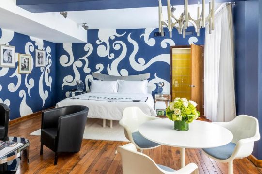 A stylish hotel room featuring a large bed with white linens against a bold blue wall with white abstract swirls, a black armchair, and a white circular table with chairs.