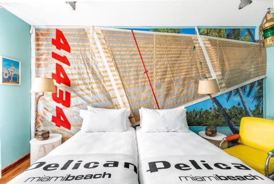 A nautical-themed bedroom with a large sail as a headboard, featuring the number "41434" and "Pelican Miami Beach" text. Two beds with white linens are flanked by lamps and a yellow chair.