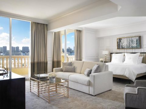 Luxurious hotel suite with a king-size bed, plush sofa, and coffee table, featuring large windows with city views and a balcony.