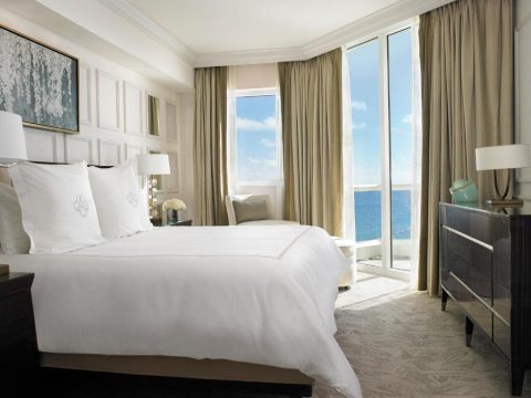 Luxurious hotel bedroom with a king-size bed, white linens, and ocean view balcony, featuring elegant drapery and a dark wood dresser.