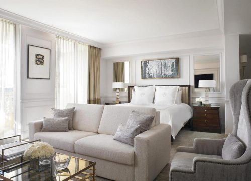 Luxurious hotel suite with a plush sofa, king-size bed with white linens, and elegant decor. A modern art piece hangs above the bed, with bedside tables and lamps adding to the sophisticated ambiance.