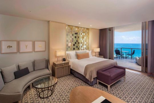 Luxurious hotel suite with a king-size bed, plush seating area, and balcony overlooking a turquoise ocean view, perfect for a beach vacation.