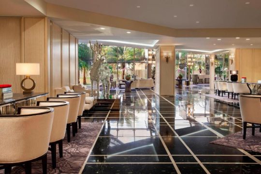 Luxurious hotel lobby with a polished black marble floor, elegant seating areas, and a large, modern white sculpture. Large windows reveal a lush outdoor garden.