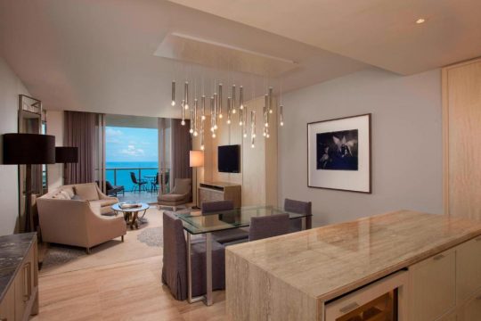 Luxurious hotel suite living room with ocean view balcony, featuring modern furniture, a dining table, and a sleek kitchen island.