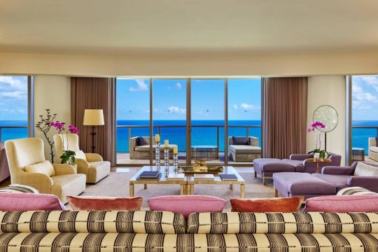 Luxurious oceanfront living room with plush purple and pink seating, facing floor-to-ceiling windows with stunning ocean views and a balcony.