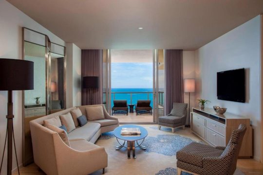 Luxurious living room with a modern sectional sofa, elegant armchairs, and a glass coffee table, opening to a balcony with ocean views and lounge chairs.