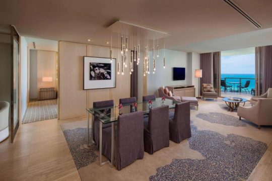 Luxury oceanfront suite with a modern dining area featuring a glass-top table and upholstered chairs, a spacious living room with plush seating, and a balcony overlooking the turquoise sea.