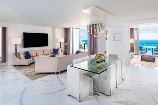 Luxurious oceanfront living room with a modern glass dining table, plush sectional sofa, and stunning turquoise ocean views from floor-to-ceiling windows.