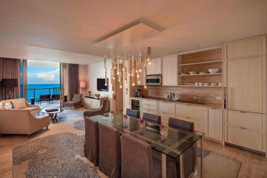 Elegant modern hotel suite dining area with a glass-top table, comfortable chairs, and a sleek kitchen with stainless steel appliances, overlooking a stunning ocean view.