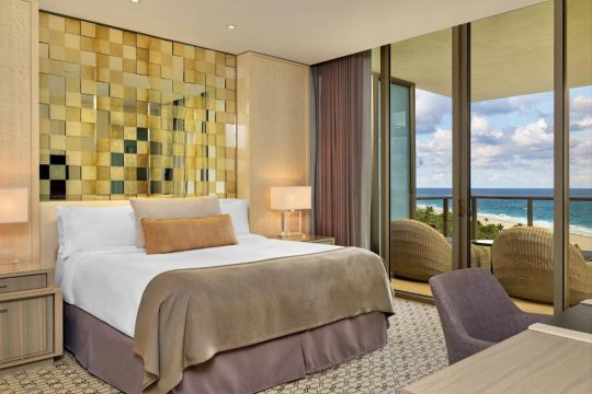 Luxurious hotel bedroom with a king-size bed, ocean view balcony, and modern decor. The room features a decorative gold and cream mosaic headboard, plush bedding, and a comfortable seating area overlooking the sea.