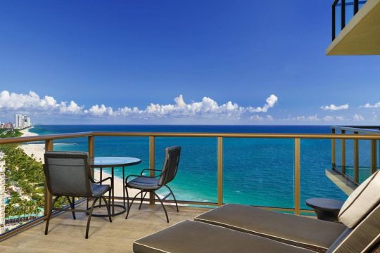 Luxury balcony with ocean view, featuring two lounge chairs, a small table, and chairs overlooking the turquoise ocean and sandy beach with palm trees.