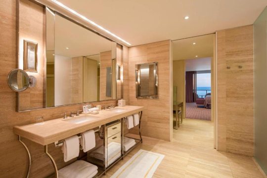 Luxurious hotel bathroom with a double vanity featuring marble countertops, modern fixtures, and ample storage for towels. The warm, natural wood tones and large mirrors create an elegant and spacious feel, leading to a glimpse of a bedroom with an ocean view.