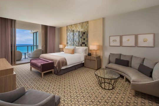 Luxurious hotel suite with a king-size bed, plush seating area, and ocean view balcony, featuring a modern design with warm tones and elegant furnishings.