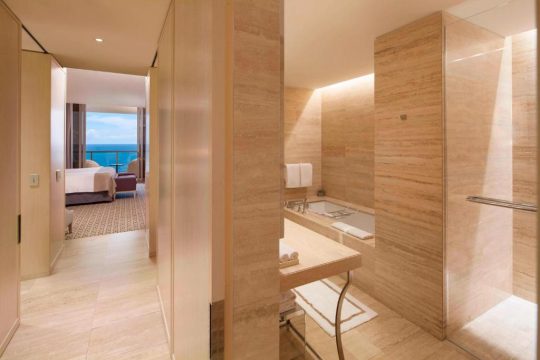 Luxurious hotel bathroom with marble walls, a deep soaking tub, and a walk-in shower, leading to a bedroom with an ocean view.