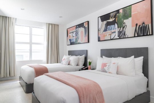 Modern hotel room with two queen beds, crisp white linens, pink accents, and abstract art on the wall.