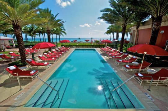 Luxury resort swimming pool with red lounge chairs and umbrellas, palm trees, and ocean view in Miami Beach.