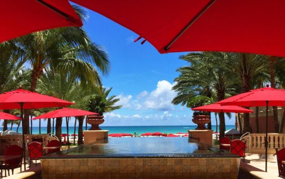 Luxury resort pool with vibrant red umbrellas and lounge chairs, surrounded by palm trees and overlooking a turquoise ocean under a bright blue sky.