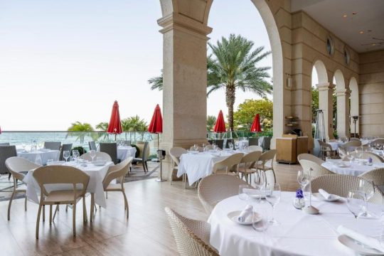 Elegant outdoor dining area with white-clothed tables, red umbrellas, and a view of the ocean, featuring palm trees and arches.