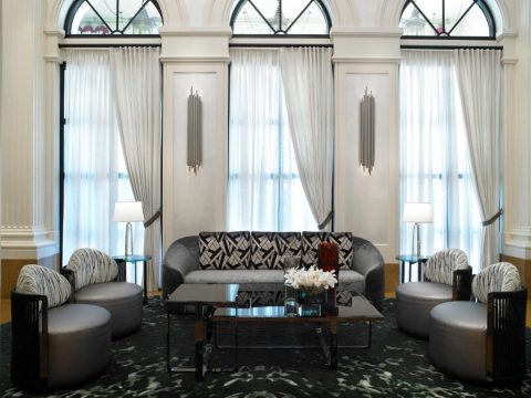 Luxurious hotel lobby seating area with a curved grey sofa, patterned cushions, and two armchairs, set against elegant white drapes and modern wall sconces.