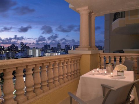 Romantic balcony dinner at dusk with city skyline view, champagne, candles, and white roses.
