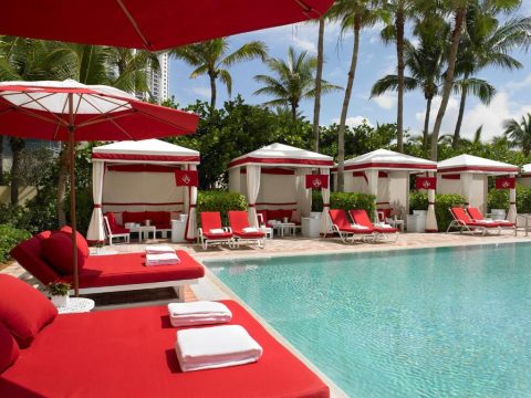 Luxurious red lounge chairs and white cabanas with red trim line a sparkling blue swimming pool at a tropical resort, with palm trees in the background.