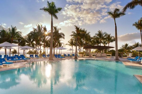 Luxury resort swimming pool with palm trees, lounge chairs, and umbrellas under a sunny sky with clouds. A swim-up bar is visible in the background.