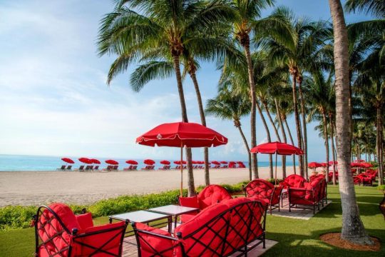 Luxury beach resort scene with vibrant red lounge chairs and umbrellas on a manicured lawn overlooking a sandy beach with palm trees and the ocean.