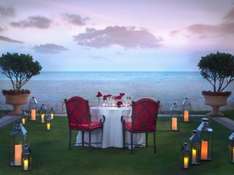 Romantic beachfront dining setup with a table set for two, adorned with roses, surrounded by glowing lanterns under a pastel sunset sky.