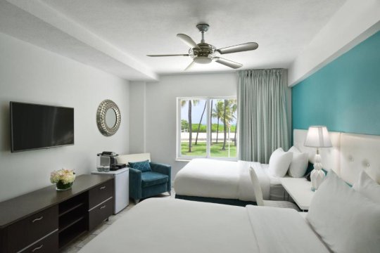 Bright hotel room with two queen beds, tropical ocean view through window, ceiling fan, and turquoise accent wall.