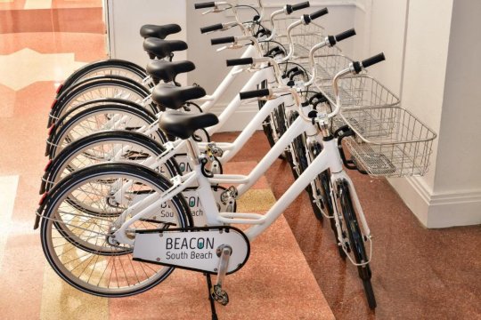 A row of white Beacon South Beach rental bikes with baskets are parked on a reddish-brown tiled floor, ready for use.