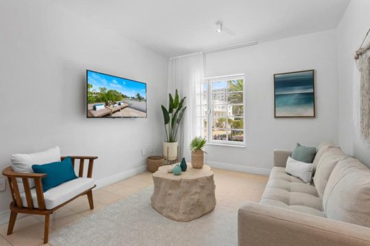 Bright and airy living room with a white sofa, armchair, natural wood accents, and a large window overlooking a sunny courtyard with palm trees.