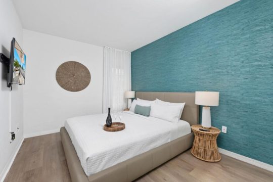 A modern bedroom with a king-size bed, white linens, a teal textured accent wall, and a wall-mounted TV.