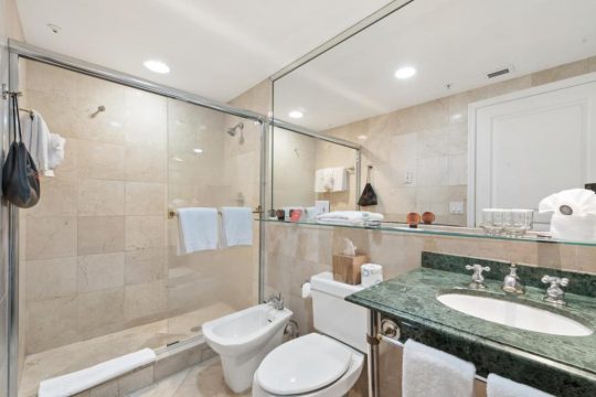 A modern bathroom features a glass-enclosed shower, a bidet, and a green marble vanity with a white sink and chrome fixtures.