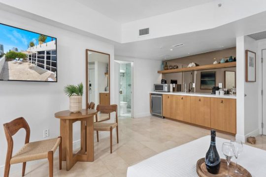 Modern studio apartment with a kitchenette, dining table, and a large TV displaying a rooftop pool scene.