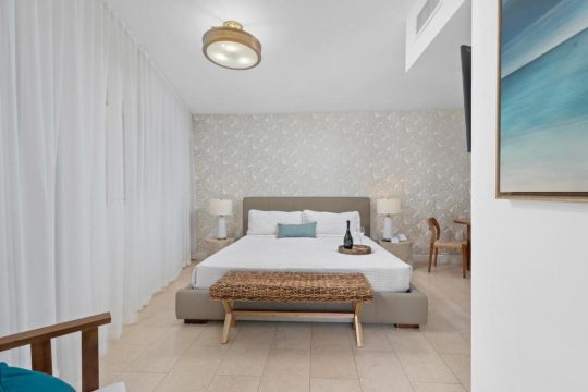 A serene hotel bedroom with a king-size bed, white linens, and a textured wallpaper. A wooden bench sits at the foot of the bed, with bedside tables and lamps on either side.