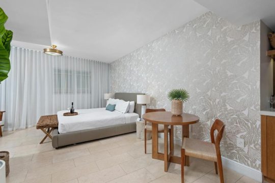 A modern studio apartment bedroom with a king-size bed, tropical leaf wallpaper, and a small dining table with chairs.