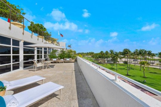 Spacious white balcony with lounge chairs overlooking palm trees and a grassy lawn, with the ocean in the distance under a bright blue sky.