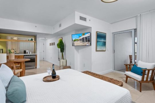 A modern studio apartment bedroom with a king-size bed, a kitchenette, and a balcony door. The room features white walls, light-tiled floors, and minimalist decor, including a large flat-screen TV mounted on the wall.