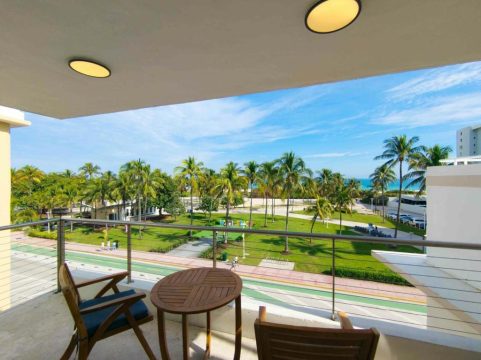 A tranquil balcony with two chairs and a small table overlooks a vibrant green park lined with palm trees and a bright blue ocean under a clear sky.