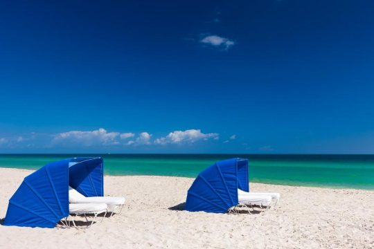 Two blue beach cabanas with white lounge chairs sit on a sandy beach, overlooking a turquoise ocean under a bright blue sky with a few wispy clouds.
