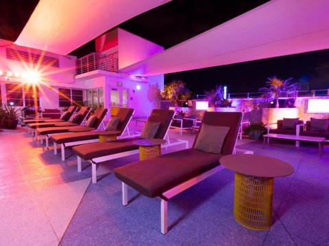 Rooftop lounge with chaise lounges and tables bathed in pink and purple light, featuring palm trees and city views at night.