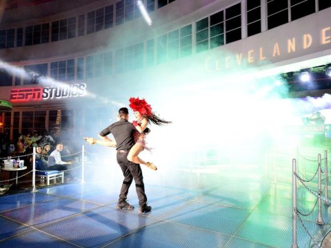 A male dancer in a black suit spins a female dancer in a red feathered costume on a bright stage in front of a crowd at ESPN Studios, Cleveland.