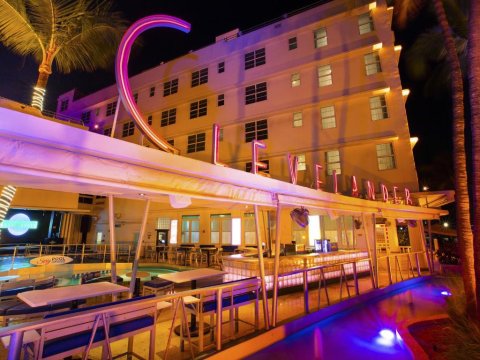 Night view of a stylish Miami hotel's outdoor pool bar and lounge area, featuring a large neon sign, palm trees, and illuminated seating.